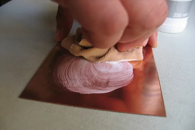 Two hands working a white cleaning paste across the copper plate with a folded cloth, a 250 ml beaker standing beside it.