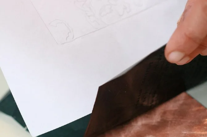 A sheet of paper lifted at one corner to show the dark carbon paper laid between it and the rocked copper plate beneath.