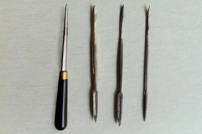 Four steel scrapers and burnishers laid side by side, one set in a black handle and three double-ended, each ground to a fine point.