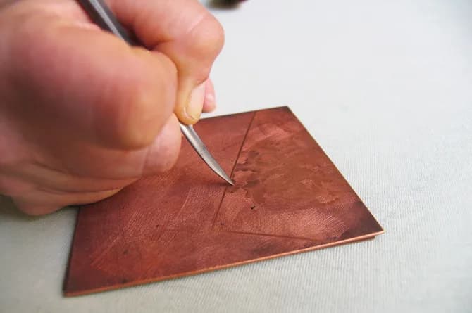 A hand drawing a fine pointed tool across the plate, a pale worked patch and a scored diagonal line already showing in the copper.